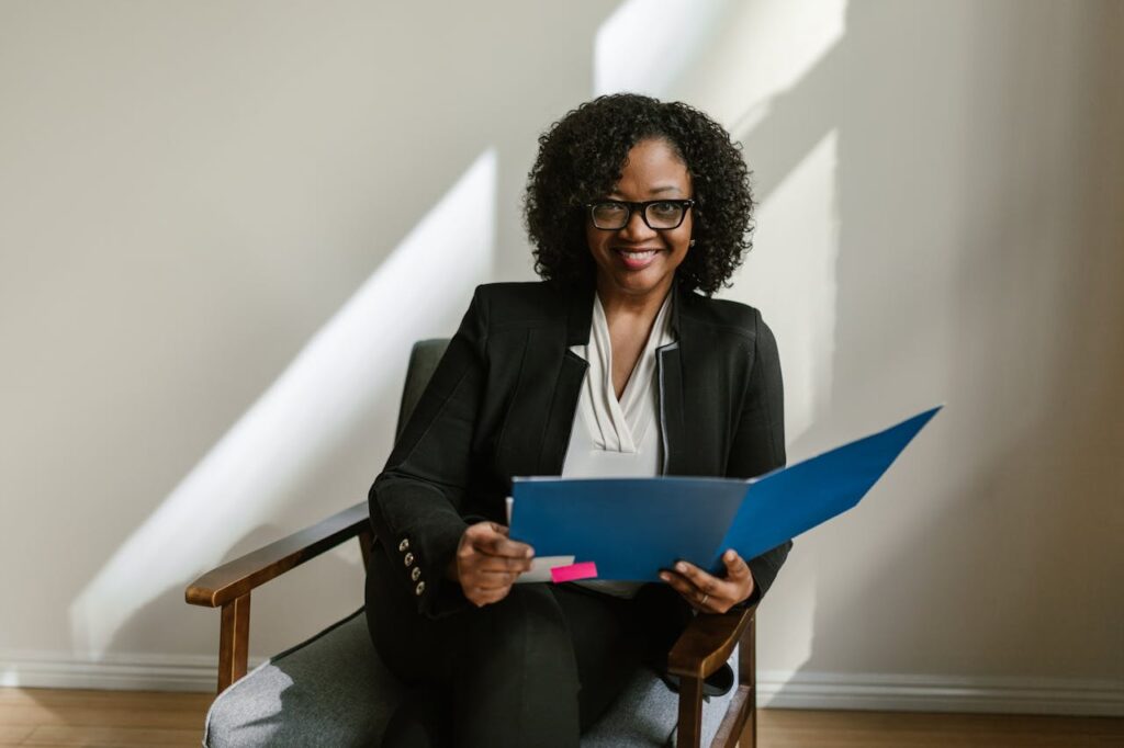Professional African American woman with glasses and curly hair holding a blue folder indoors.
